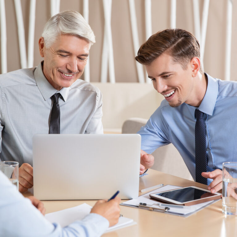 Smiling senior and young businessmen and female colleague working, discussing ideas and sitting around small table with laptop in cafe. Men are using laptop. Woman is writing and seen partly.